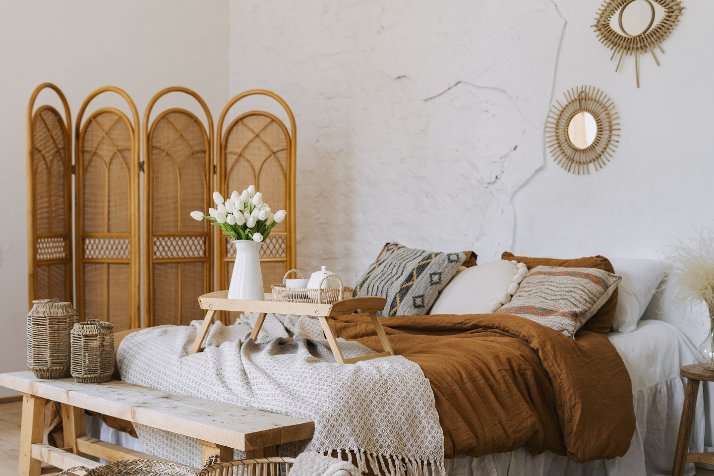 Comfort bedroom in boho style interior with pillow and plaid on bed, wooden bench seat, bamboo dressing screen, home decor. Breakfast tray with flowers and teapot.