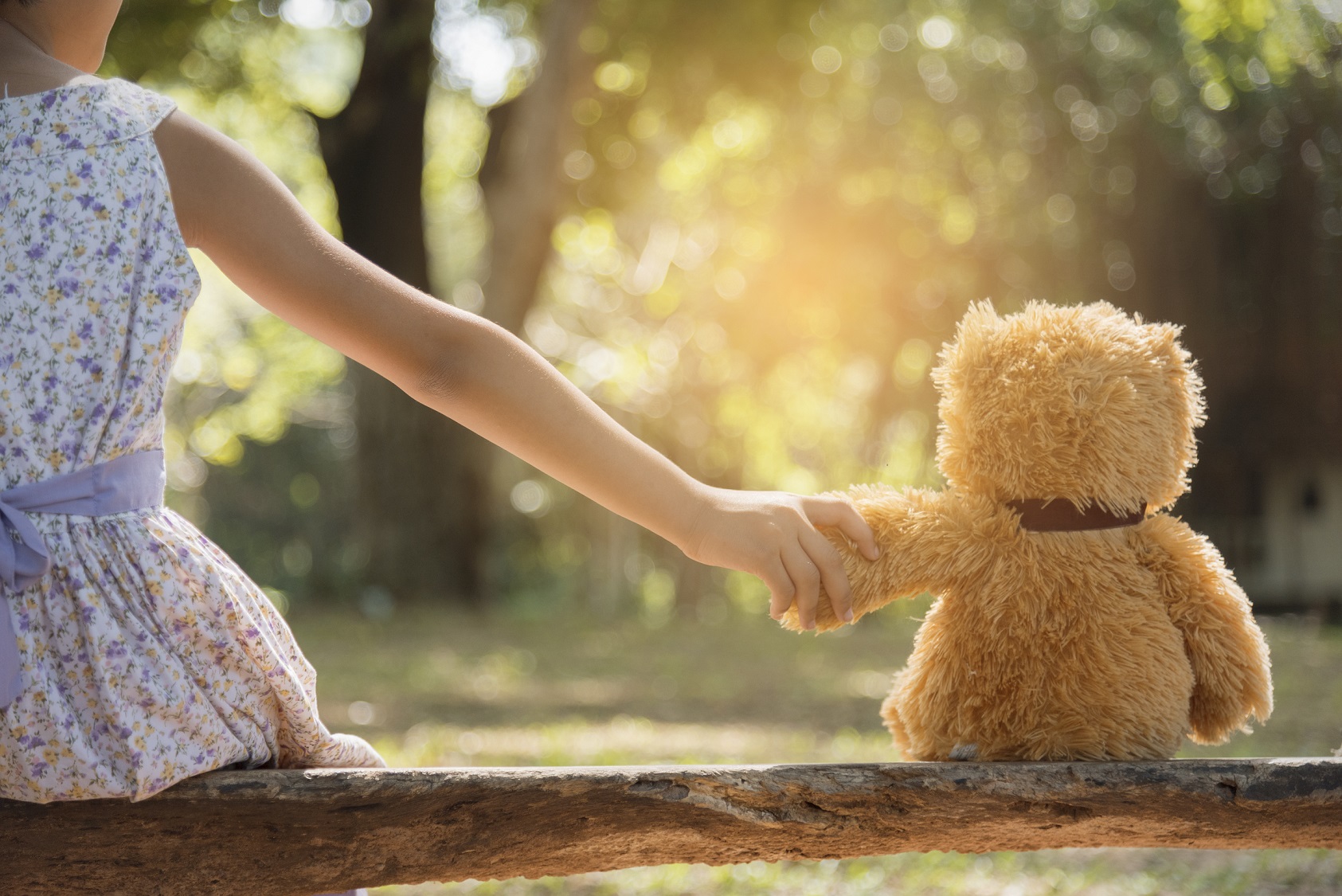 A girl holding hands with a teddy bear sitting on a wooden bench